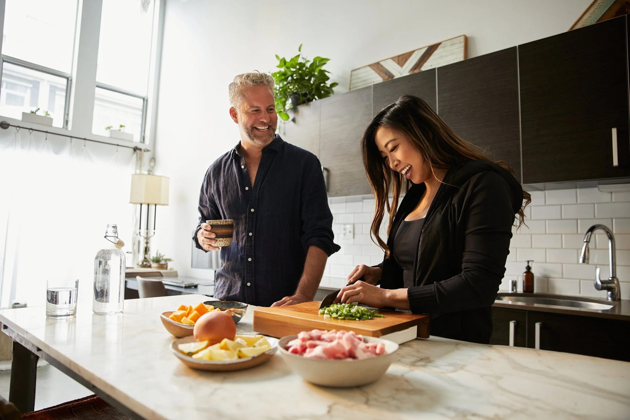 A man and woman laughing and preparing food in a modern kitchen with natural light
